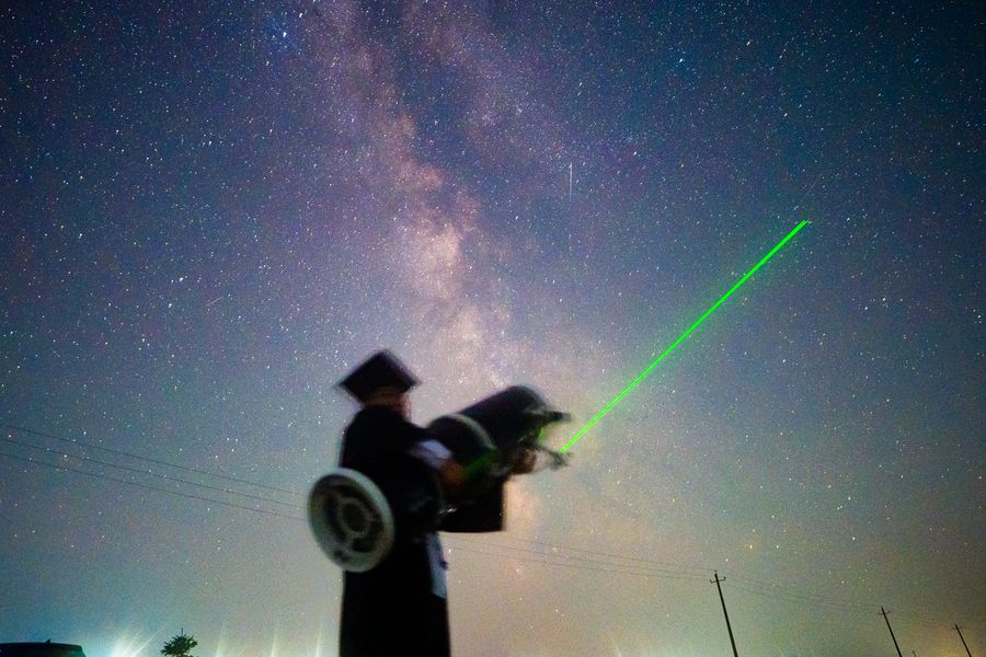 绿色光柱斜着划过星空，一位毕业生低头站在画面中间，旁边还有人蹲着