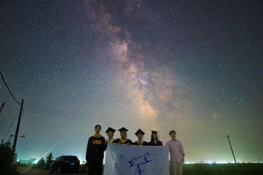 一群毕业生在轿车旁合影，手里举着蓝色横幅，头顶是横贯夜空的银河