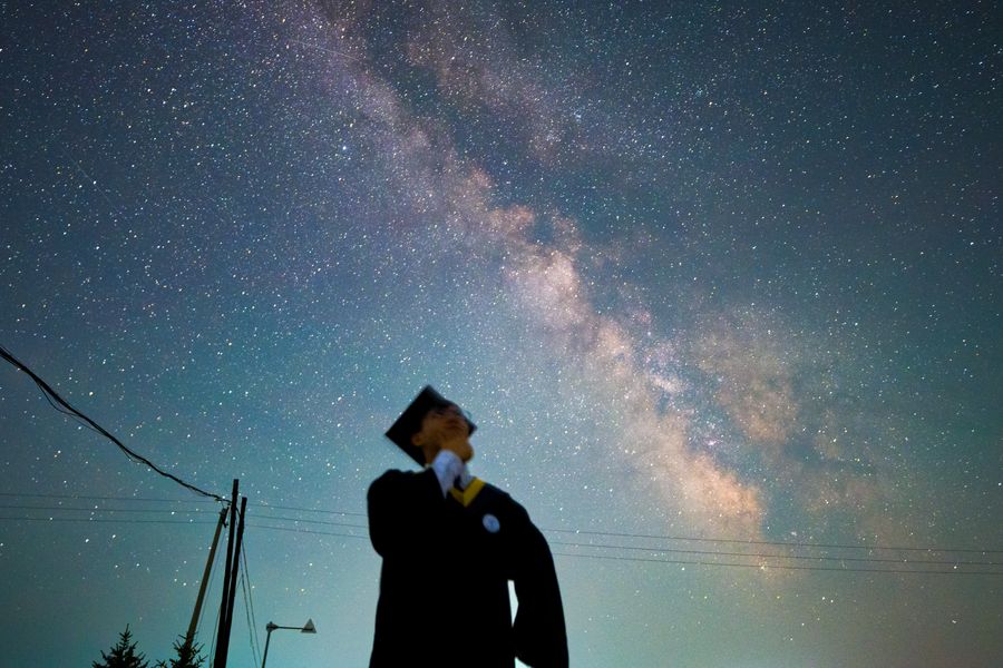 穿学士服的毕业生仰头望向银河，银河从头顶斜贯而过，背景是夜空与远处的屋脊
