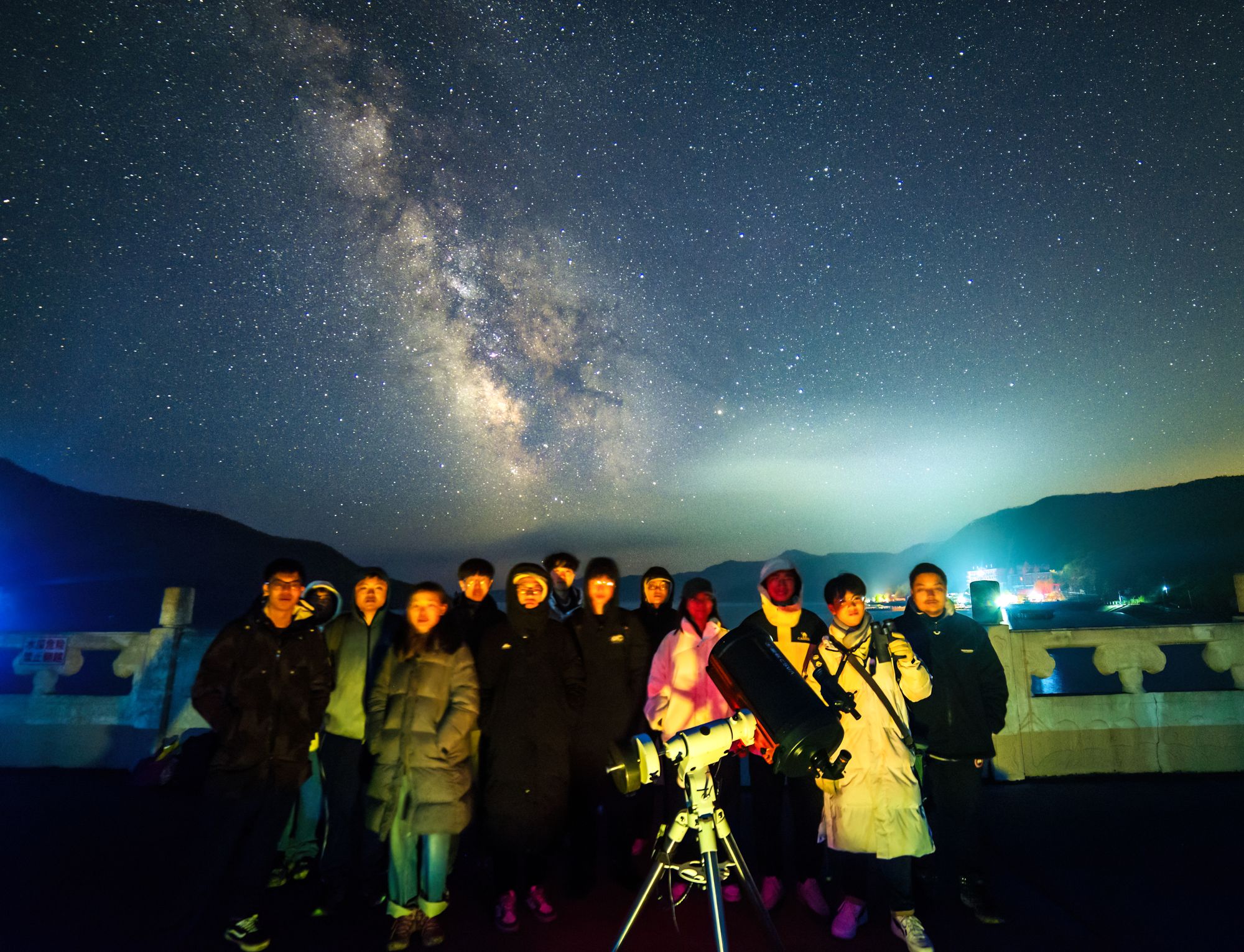 十几位社团成员在星星哨水库边与一台望远镜合影，头顶是银河