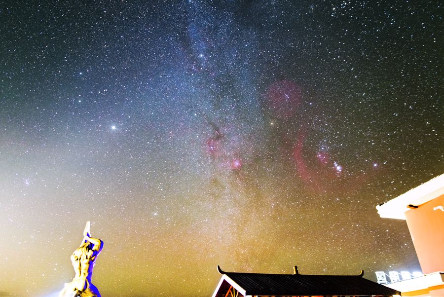 银河横贯夜空，下方亮着两盏灯，地面与树影被照出轮廓，低空有一片绿光