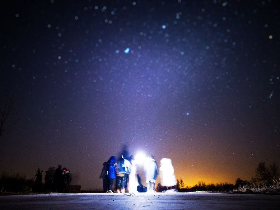 新山水库的星空下，四五个人背对镜头站着，中间一盏强光把人与前景照得发白、边缘晕开，天上的星点虚成柔光，右侧地平线留着一层橙色的光