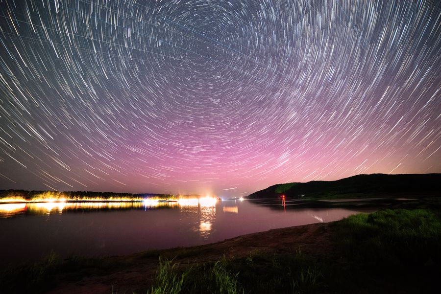 星轨在江面上方绕成半圈，左下是岸边温暖的灯光，水面映出天空的颜色